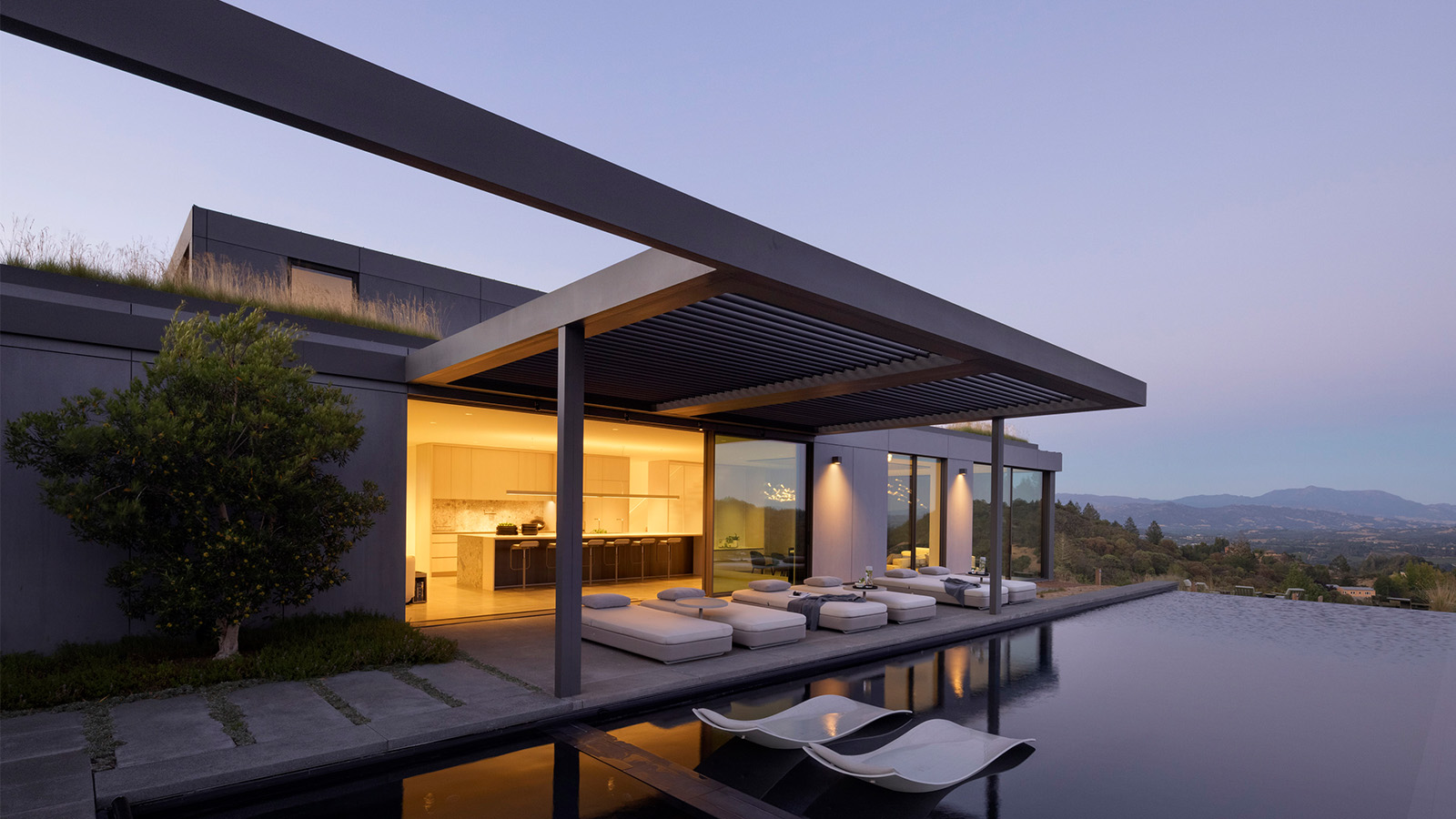 A beautiful modern home with a cantilevered pergola, glass-walled kitchen, and infinity pool